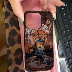 Puede incluir: Funda de tel&eacute;fono negra con un collage de un jugador de f&uacute;tbol en acci&oacute;n. La funda tiene un borde negro y un dise&ntilde;o con el logo del equipo y la imagen del jugador. El fondo es una mezcla de tonos marrones y naranjas.