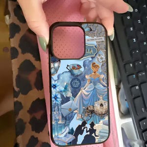 May include: A black phone case featuring a collage of Cinderella-themed imagery. The design includes Cinderella in a blue gown, a carriage, and the text "SO THIS IS LOVE." Additional elements include a teacup, flowers, and a castle.