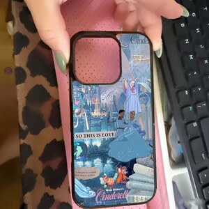 May include: A phone case featuring a Cinderella-themed design. The case has a black border and a colorful illustration of Cinderella, the castle, and the text "SO THIS IS LOVE" and "Walt Disney's Cinderella". The background is a light blue and pink.