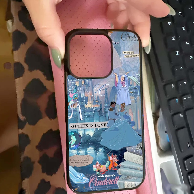 May include: A phone case featuring a Cinderella-themed design. The case has a black border and a colorful illustration of Cinderella, the castle, and the text "SO THIS IS LOVE" and "Walt Disney's Cinderella". The background is a light blue and pink.