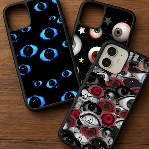 Pode incluir: Três capas de telefone pretas com designs de olhos. Uma capa apresenta olhos azuis brilhantes em um fundo preto. Outra tem vários globos oculares com estrelas. A terceira capa é coberta por uma colagem de olhos diferentes.
