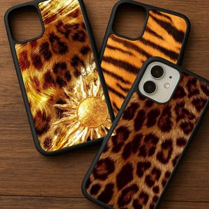 May include: Three phone cases with black borders. One case features a gold sun design on a leopard print background. Another has a tiger stripe pattern, and the third has a leopard print design. The cases are on a wooden surface.