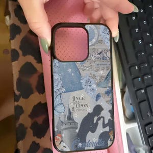 May include: Black phone case with a Cinderella theme. The case features a collage of blue floral patterns, musical notes, and silhouettes of Cinderella and Prince Charming. Text includes "Once upon a time..." and "Cinderella."