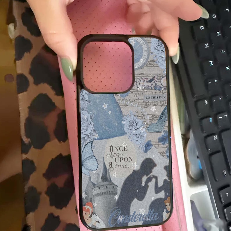May include: Black phone case with a Cinderella theme. The case features a collage of blue floral patterns, musical notes, and silhouettes of Cinderella and Prince Charming. Text includes "Once upon a time..." and "Cinderella."