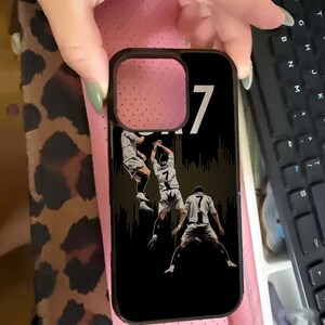 Puede incluir: Funda de tel&eacute;fono negra con un gr&aacute;fico de jugador de f&uacute;tbol. La funda tiene un borde negro y un fondo negro con un gr&aacute;fico de un jugador de f&uacute;tbol con uniforme blanco y gris. El n&uacute;mero 7 se muestra de forma destacada.