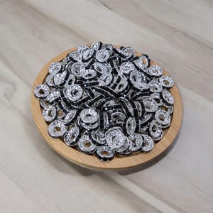 May include: A bowl filled with silver and black round beads with rhinestones. The beads have a small hole in the centre.