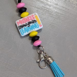 May include: A white rectangular charm with the text "Mistakes show you're learning" and a yellow pencil illustration. The charm is attached to a black cord with pink, black, and yellow beads. A silver clip and a light blue tassel are attached to the bottom of the cord.