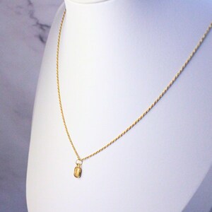 May include: A gold-colored necklace with a small pendant. The pendant is rectangular with etched details. The necklace is displayed on a white bust against a white and gray background. The chain is delicate and twisted.
