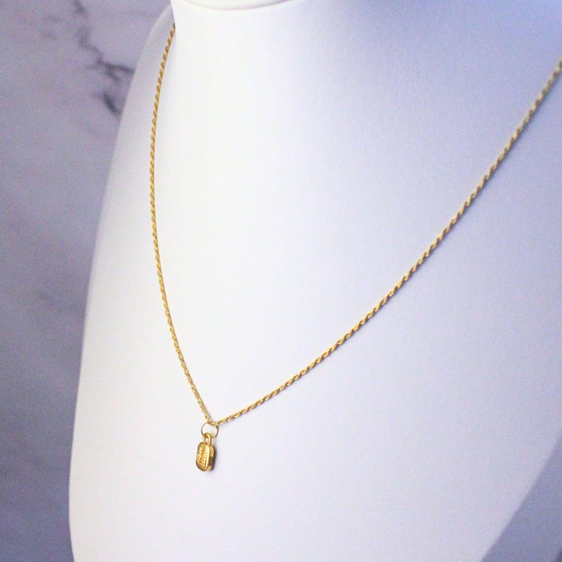 May include: A gold-colored necklace with a small pendant. The pendant is rectangular with etched details. The necklace is displayed on a white bust against a white and gray background. The chain is delicate and twisted.
