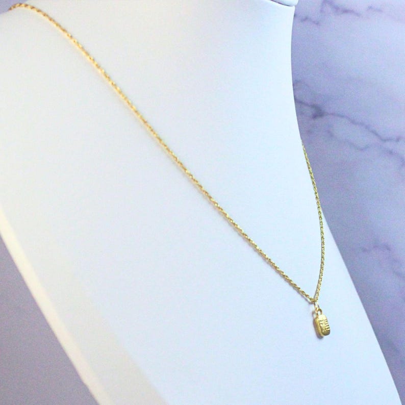 May include: A gold-colored necklace with a small rectangular pendant. The pendant has etched details and hangs from a delicate chain. The necklace is displayed on a white mannequin bust against a soft, marbled background.
