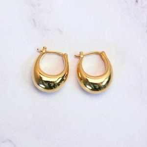 May include: A pair of gold-colored hoop earrings. The earrings have a smooth, polished surface and a rounded, teardrop shape. The earrings are displayed on a white marble surface.