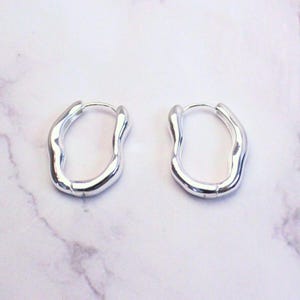 May include: A pair of silver-coloured, organic-shaped hoop earrings. The earrings have a smooth, reflective surface and a unique, irregular form. The hoops are designed to hug the earlobe. The earrings are displayed on a white and grey marble surface.