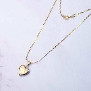 Può includere: Una collana color oro caratterizzata da una delicata catena intrecciata e da un ciondolo a forma di cuore liscio e lucido. La collana include un piccolo meccanismo di chiusura circolare vicino alla parte superiore, con una finitura metallica semplice e riflettente.