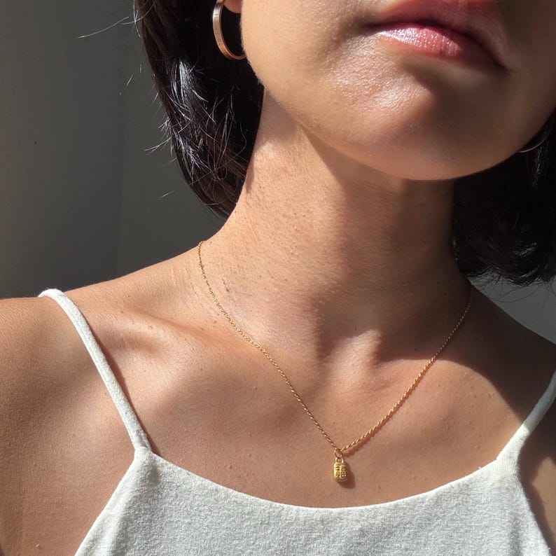 May include: A gold-colored necklace with a small, rectangular pendant. The necklace is delicate and hangs on a person's neck, paired with gold hoop earrings and a white tank top. The jewelry is simple and elegant.