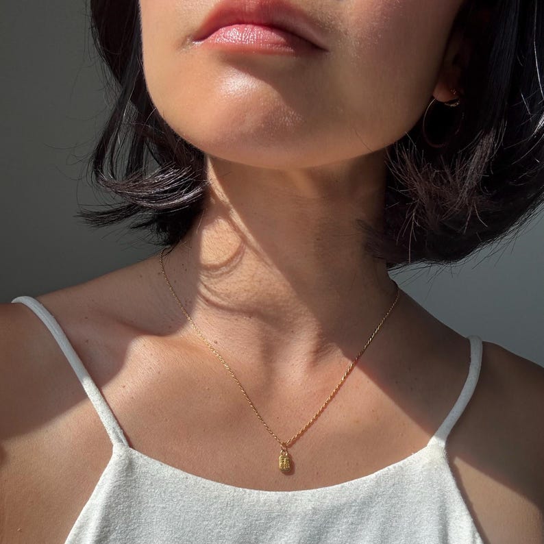 May include: A delicate gold necklace with a small pendant. The pendant has a textured design. The necklace is worn on a person wearing a white tank top. The image is well-lit, highlighting the jewelry.