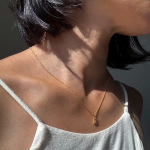May include: A delicate gold necklace with a small rectangular pendant. The pendant has a textured surface and is suspended from a fine chain. The necklace is worn against a white tank top.