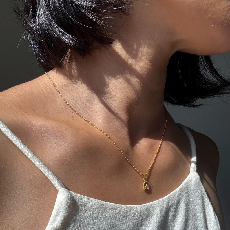 May include: A delicate gold necklace with a small rectangular pendant. The pendant has a textured surface and is suspended from a fine chain. The necklace is worn against a white tank top.