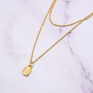 May include: A gold-colored, layered necklace with a small rectangular pendant. The pendant has Chinese characters engraved on it. The necklace is displayed on a white and gray marble surface.