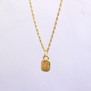 May include: A gold-colored necklace with a rectangular pendant. The pendant features an embossed design, and the chain has a twisted rope style. The necklace is displayed against a white background.