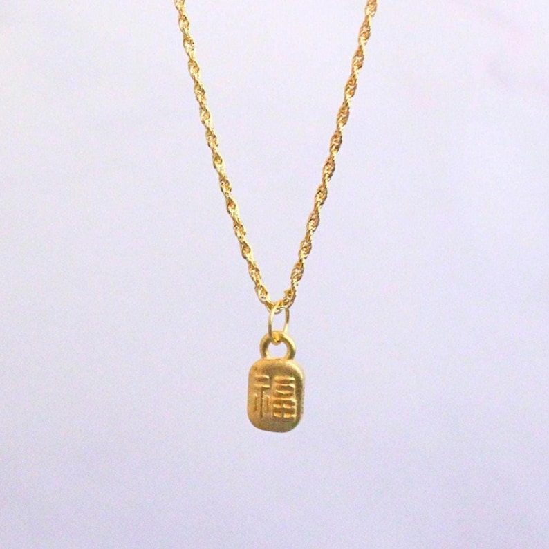 May include: A gold-colored necklace with a rectangular pendant. The pendant features an embossed design, and the chain has a twisted rope style. The necklace is displayed against a white background.