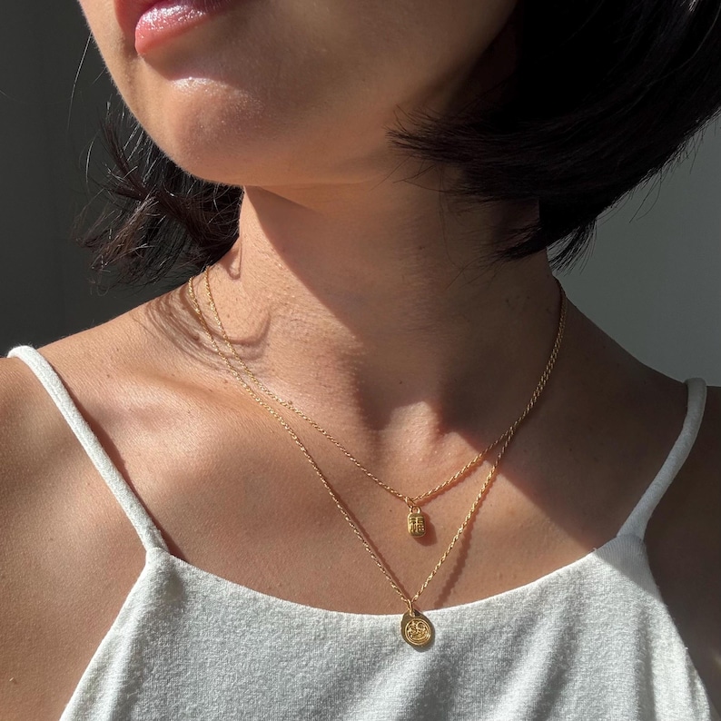 May include: A layered gold necklace set. The top necklace features a small, textured pendant, while the lower necklace has a circular pendant with a design. The necklaces are worn over a white tank top.