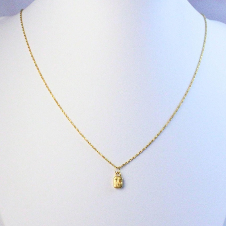 May include: A delicate gold-colored necklace with a small rectangular pendant. The pendant has an engraved design. The necklace is displayed on a white background.