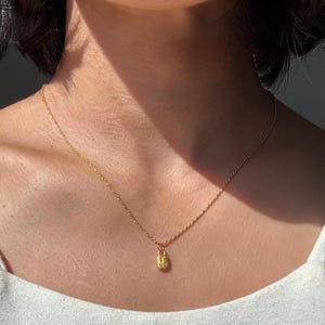 May include: A gold-colored necklace with a small rectangular pendant. The pendant has an embossed design. The necklace chain is delicate and hangs on a person's neck.