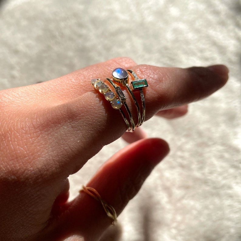May include: A set of four gold rings with gemstones. The rings feature a moonstone, an emerald, and a row of opals. The rings are stacked on a finger.