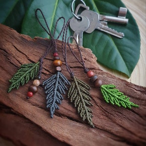 May include: Four handmade macrame leaf keychains with wooden beads in shades of green, brown, and red. The keychains are hanging from a wooden surface.