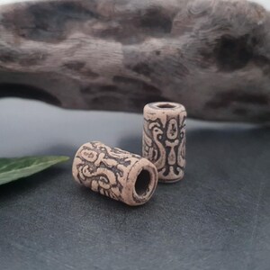 May include: Two beige ceramic beads with intricate black designs. The beads are cylindrical and have a smooth, polished finish.