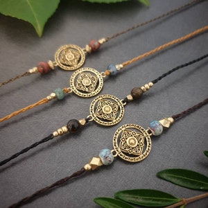 Könnte beinhalten: Vier handgefertigte Armbänder mit filigranen goldfarbenen runden Anhängern und verschiedenen Perlen. Die Armbänder sind auf braunen, beigen und schwarzen Kordeln aufgefädelt. Die Anhänger haben ein detailliertes Blumenmuster. Dunkelgrauer Hintergrund.
