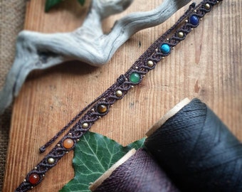 Könnte beinhalten: Ein braunes Lederarmband mit sieben bunten Edelsteinen und goldenen Perlen. Das Armband liegt auf einer Holzoberfläche mit Spulen braunen Fadens.