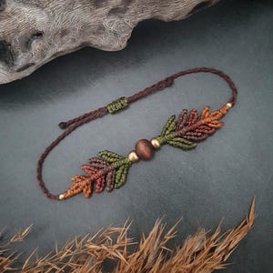 May include: A macrame bracelet with leaf motifs in green, brown, and orange. The bracelet has a wooden bead and gold-coloured accents. The cord is dark brown. The bracelet is likely to fit a wrist size of approximately 15-20 cm.