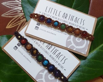 Könnte beinhalten: Zwei braune Karton-Etiketten mit weißem Text, der "LITTLE ARTIFACTS Schmuck & Dread-Accessoires inspiriert von der Natur" lautet und Kontaktinformationen enthält. Jedes Etikett hat ein braunes und goldenes Armband mit bunten Edelsteinperlen.