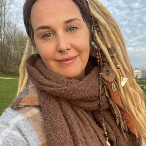 Op de afbeelding: Een vrouw met blonde dreadlocks draagt een bruine gebreide sjaal en een bruin hoofdband. Ze draagt een bruin en blauw geruit jasje. De dreadlocks hebben kralen en veren.