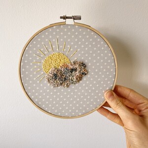 May include: Embroidered hoop art featuring a yellow sun with radiating lines and a textured cloud. The design is on a gray fabric with a white cross pattern, set within a wooden hoop. A unique piece of wall decor.
