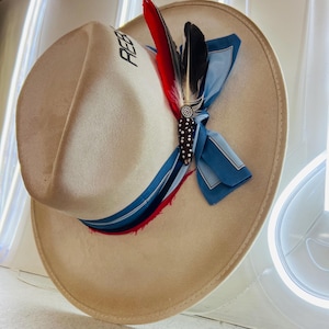 May include: A beige wide-brimmed hat featuring the text REB in black lettering on the crown. The hat is adorned with a blue silk ribbon, a red feather, a black and white feather, and a decorative brown beaded accent with silver studs.