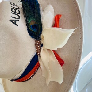 Puede incluir: Un sombrero de ala ancha color crema con la palabra "AUBURN" bordada en negro. El sombrero est&aacute; adornado con una pluma de pavo real, una flor de tela blanca y una cinta en azul, naranja y blanco. Un sombrero decorativo para eventos.