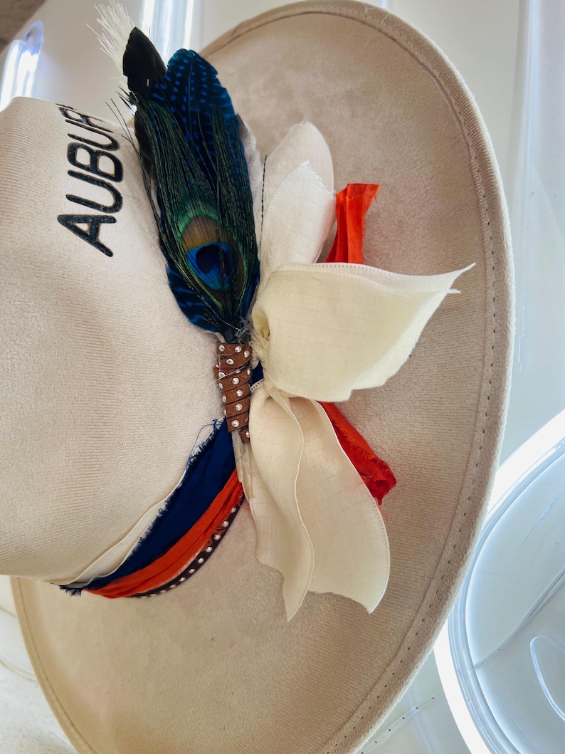 Puede incluir: Un sombrero de ala ancha color crema con la palabra "AUBURN" bordada en negro. El sombrero est&aacute; adornado con una pluma de pavo real, una flor de tela blanca y una cinta en azul, naranja y blanco. Un sombrero decorativo para eventos.