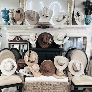 Puede incluir: Una colección de sombreros de vaquero de varios colores y estilos, expuestos en una estantería y sillas. Los sombreros presentan elementos decorativos como plumas y cintas. La paleta de colores incluye marrón, blanco y beige.