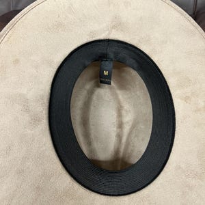 Puede incluir: Sombrero fedora beige con una banda interior negra. El sombrero es de talla mediana, indicado por la etiqueta "M". El ala ancha y el material sugieren una textura similar a la gamuza. Un accesorio de moda.