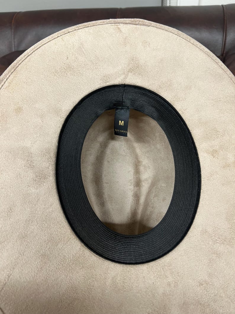 Puede incluir: Sombrero fedora beige con una banda interior negra. El sombrero es de talla mediana, indicado por la etiqueta "M". El ala ancha y el material sugieren una textura similar a la gamuza. Un accesorio de moda.