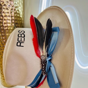 May include: A beige hat with the word "REBS" printed in black. The hat is decorated with red, black, and white feathers, a blue scarf, and a decorative jeweled pin. The hat has a wide brim and a soft texture.