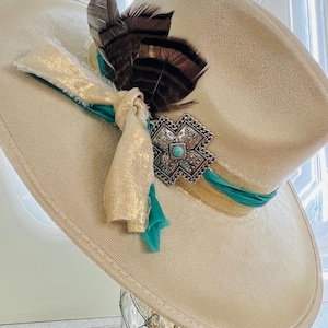 May include: A cream-coloured cowboy hat with a decorative turquoise and silver-tone metal embellishment. The hat features a turquoise ribbon, gold fabric, and brown and black feathers. A stylish western accessory.