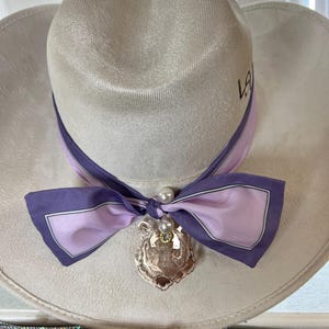 May include: A light gray cowboy hat with a decorative purple and pink scarf tied around the base. A gold-colored tiger pendant with a pearl and rhinestone accents hangs from the scarf. The hat has the letters "LA" on the side.