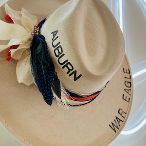 Puede incluir: Sombrero de vaquero color crema con la palabra "AUBURN" bordada en negro. El sombrero est&aacute; decorado con una cinta azul, naranja y blanca, plumas y una flor blanca. Las palabras "WAR EAGLE" est&aacute;n bordadas en el ala.