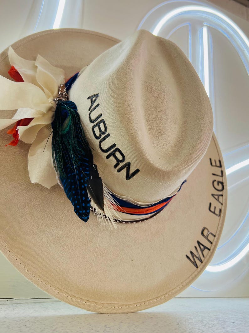 Puede incluir: Sombrero de vaquero color crema con la palabra "AUBURN" bordada en negro. El sombrero est&aacute; decorado con una cinta azul, naranja y blanca, plumas y una flor blanca. Las palabras "WAR EAGLE" est&aacute;n bordadas en el ala.