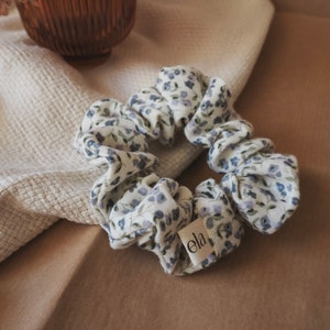 Könnte beinhalten: Ein weißer Haargummi mit einem kleinen Blumenmuster in Blau- und Lilatönen. Der Haargummi hat ein kleines Etikett mit der Aufschrift "ela" darauf.