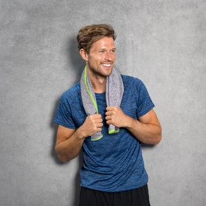 May include: A man wearing a blue athletic shirt with a gray and green towel around his neck. He is smiling and looking to the right.