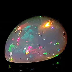 May include: A pear-shaped opal gemstone with a milky white base. The stone displays vibrant flashes of color, including green, pink, and orange, against a dark background. The opal is polished and smooth, showcasing its natural beauty.